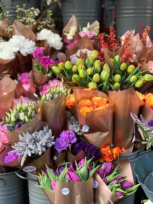 An assortment of fresh flowers arranged in brown kraft paper wrapping and displayed in metal buckets. The bouquet includes closed green tulips, orange and pink roses, purple lisianthus, white astilbes