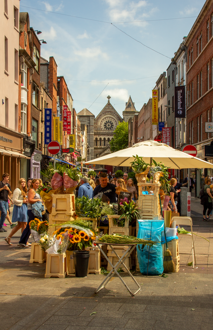 A bustling street scene on St Johns Wood High Street with an outdoor flower stall featuring fresh, vibrant arrangements of sunflowers, pink roses, and greenery in wooden crates and vases. The stall is