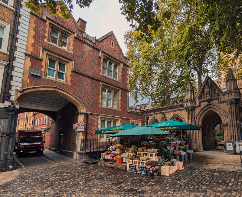 A flower stall located near a historic stone archway and red-brick building in St Johns Wood, featuring an array of fresh flowers arranged in wooden crates and bouquets, under a green umbrella with ye