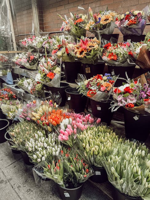 A display of fresh floral bouquets arranged in black buckets at a flower shop, showcasing a variety of flowers including pink and white tulips, orange and pink roses, and mixed seasonal blooms wrapped
